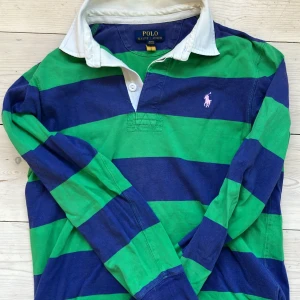 Randig pikétröja från Ralph Lauren - Snygg pikétröja från Ralph Lauren med breda gröna och blå ränder, vit krage och klassisk rosa logga på bröstet. Långärmad modell i mjuk bomull. 