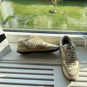 Valentino Garavani Rockrunner sneakers - Beiga Valentino Garavani Rockrunner sneakers med ikoniska nitar på sidorna. Skorna har en mix av mocka och textil, snörning och svart mönstrad sula. Snygg och lyxig design som sticker ut med sina detaljer och den klassiska Valentino-looken.