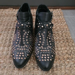 Svarta boots med nitar från K.Cobler - Svarta boots i skinn från K.Cobler med coola guldfärgade nitar och öglor över hela skon. Spetsig tå, låg klack och snygg spänndetalj vid ankeln. Dragkedja på insidan för enkel påtagning. Perfekta för dig som vill sticka ut med din stil.