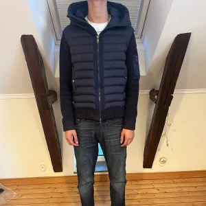 Mörkblå Moncler pufferjacka med huva - Säljer en mörkblå pufferjacka från Moncler med stickade ärmar och vadderad front. Jackan har huva, dragkedja framtill och en ficka med dragkedja på ärmen. Klassisk Moncler-logga på vänster arm och snygga ribbade muddar.