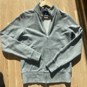 Grå ziptröja från Gant - Säljer en grå ziptröja från Gant i storlek S. Tröjan har hel dragkedja, ståkrage och två fickor framtill. Liten Gant-logga broderad på bröstet. Mjuk och skön bomullsblandning, perfekt för lager-på-lager. Klassisk och stilren design.