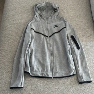 Grå hoodie från Nike med dragkedja - Grå hoodie från Nike med svart logga på bröstet och svarta detaljer. Tröjan har hel dragkedja framtill, huva och en praktisk ficka med dragkedja på ena ärmen. Tillverkad i mjuk bomullsmix, perfekt för chill eller träning. Svarta kantband vid ärmslut och nederkant.