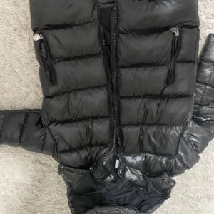 Svart Moncler pufferjacka med huva - Säljer en svart pufferjacka från Moncler med glansig finish och klassisk logga på ärmen. Jackan har dragkedjor på fickorna, huva och quiltad design. Perfekt för kalla dagar och riktigt snygg streetstil. Storleken är M men passar även L