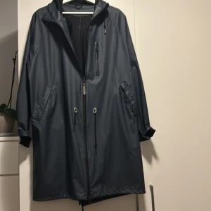Mörkblå regnjacka från Stenk L-XL - Snygg mörkblå regnjacka från Stenk i storlek L-XL. Jackan har huva, dragkedja framtill, snörning i midjan och flera fickor. Materialet är syntet och jackan har en loose passform med långa ärmar. Perfekt för regniga dagar. knappt använd