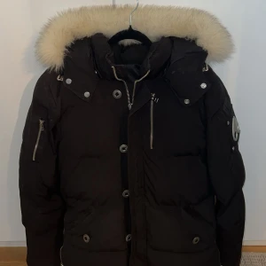 Svart dunjacka Moose Knuckles med päls - Säljer en svart dunjacka från Moose Knuckles med fluffig vit päls runt huvan. Jackan har flera dragkedjor, knappar och en ikonisk logga på ärmen. Perfekt för kalla vinterdagar och riktigt snygg streetstil.