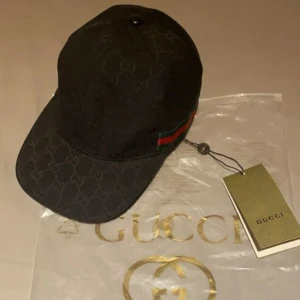 Gucci Keps - Exklusiv svart keps från Gucci med klassiskt GG-monogram över hela kepsen och ikoniska gröna och röda ränder på sidan. Justerbar rem baktill i skinn och tillverkad i Italien. Perfekt accessoar för dig som vill sticka ut med lyxig streetstyle.