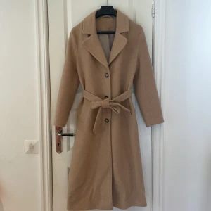 INTRESSEKOLL Beige kappa i ull från & Other Stories - INTRESSEKOLL! Snygg beige kappa från & Other Stories Paris Atelier, tillverkad i Pontetorto-tyg från Italien. Klassisk rak modell med knappar framtill, två fickor och ett bälte i midjan. Långärmad och med krage, perfekt för höst och vinter. Endast använd ett fåtal gånger.