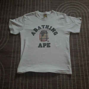 Vit A Bathing Ape t-shirt M - Vit t-shirt från A Bathing Ape med klassiskt tryck på bröstet och rutigt ape-head motiv. Skön passform och crewneck. Ikonisk logga på ärmen och detaljerad etikett. Perfekt för dig som gillar streetwear och vill sticka ut.