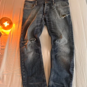 Blå slitna jeans från Nudie - Pris kan diskuteras. Säljer ett par blå jeans från Nudie med snygga slitningar och hål på låret och knät. Klassisk femficksmodell med orange sömmar och Nudie-logga på bakfickan. Jeansen har en rak passform och är tillverkade i mjukt denimtyg. Slit längst ner.