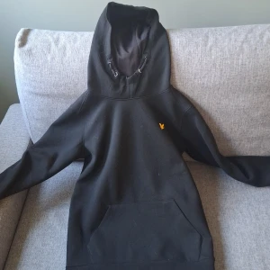 Svart hoodie från Lyle & Scott - Svart hoodie från Lyle & Scott med klassisk gul logga på bröstet. Tröjan har stor huva med snörning och en rymlig magficka. Tillverkad i mjuk polyesterblandning med lite stretch för extra komfort. Perfekt för chill dagar eller när du vill ha en enkel och stilren look. Tröjan är ganska sparsamt använd och är mycket bekväm.