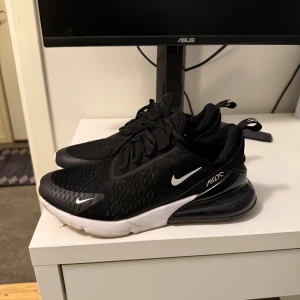 Nike Air Max 270 svart, vita sneakers - Svarta Nike Air Max 270 sneakers med vit sula och tydlig Air Max-bubbla i hälen. Skorna har mesh-överdel för bra andningsförmåga, snörning och diskreta vita detaljer som swoosh-loggan. Perfekta för dig som gillar sportig och modern stil.