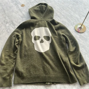 Olivgrön hoodie från Zadig & Voltaire - Cool olivgrön hoodie från Zadig & Voltaire med dragkedja och huva. Storlek S-Xs. Baksidan har en stor vit döskalle som ger en edgy vibe. Tröjan är stickad och har två fickor framtill. Perfekt för dig som vill sticka ut med stil. Passar mig som är runt 177. Priset är inte hugget i sten. Några funderingar är de bara att skriva!
