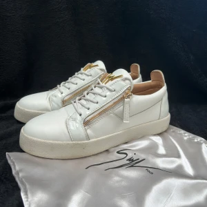 Vita sneakers från Giuseppe Zanotti - Snygga vita sneakers från Giuseppe Zanotti med guldfärgade dragkedjor på sidorna och ikonisk logga på plösen. Skorna är i skinn och har snörning samt platt sula. Perfekta för dig som gillar exklusiv streetstyle med lyxiga detaljer.