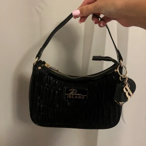 Svart handväska från River Island - Snygg svart handväska från River Island med glansig finish och quiltad struktur. Väskan har guldfärgade detaljer, dragkedja upptill och en dekorativ nyckelring med RI-logga. Kommer med både kort och lång axelrem för olika stilar.