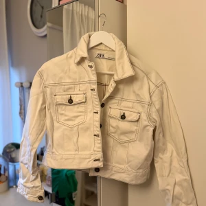 Vit jeansjacka från ZARA - Snygg vit jeansjacka från ZARA med kontrastsömmar och bruna knappar. Jackan har två bröstfickor med lock och knapp, klassisk krage och croppad passform. Perfekt för dig som vill ha en stilren och trendig look.