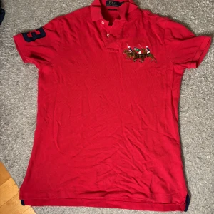Röd pikétröja från Polo Ralph Lauren - Röd pikétröja från Polo Ralph Lauren med broderat polospel-motiv på bröstet och en stor blå siffra 3 på ärmen. Klassisk krage med knappar och korta ärmar. Perfekt för en sportig och stilren look.