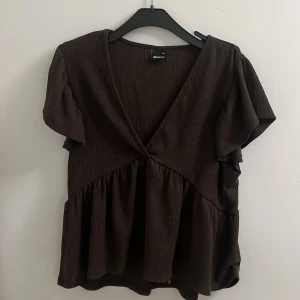 Brun omlottblus från Gina Tricot - Supersöt brun blus från Gina Tricot med omlott-look och v-ringning. Toppen har korta, voluminösa ärmar och ett rynkat, flowy parti nedtill. Materialet är lätt strukturerat och ger en snygg textur. Perfekt att styla med jeans eller kjol för en trendig look.