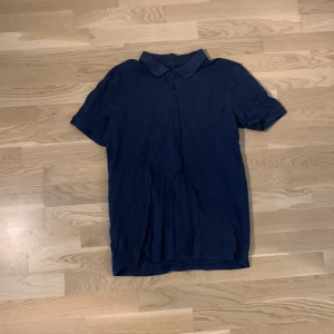 Mörkblå pikéskjorta från Newbody - Snygg mörkblå pikéskjorta från Newbody i storlek M. Skjortan har klassisk krage, korta ärmar och knappar framtill. Perfekt för dig som gillar enkel och stilren look.