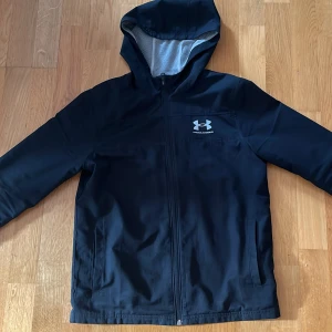 Under Armour vindjacka - Svart vindjacka från Under Armour med huva och dragkedja framtill. Jackan har två sidofickor och logga på bröstet. Perfekt för blåsiga dagar och sportiga outfits. Det står någon konstig storlek i men passar mig som är 165 så motsvarar nog XS, skick 8/10 (ett litet hål i fickan)