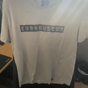 Vit ODZ t-shirt Zubbkultur Storlek: S - Vit t-shirt från Dunderkläder med svart tryck där det står 'ZUBBKULTUR' på bröstet. Klassisk passform och rund halsringning, perfekt för dig som gillar streetwear och vill sticka ut med ett unikt tryck. Storlek: S