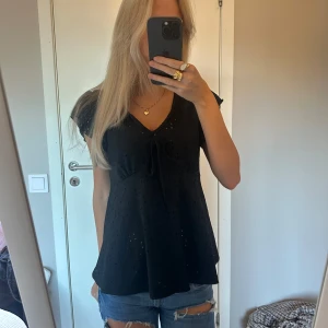 Svart blus med volangärm och knyt - Säljer en svart blus med v-ringning, knyt framtill och volangärmar. Blusen har broderade hål som ger en luftig känsla och ett snyggt mönster. 