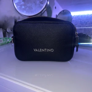 Svart axelväska från Valentino - Snygg svart axelväska från Valentino med stilren, fyrkantig form och guldfärgade dragkedjor. Väskan är tillverkad i strukturerat skinnliknande material och har ett fack med dragkedja på baksidan samt flera innerfack för smart organisering. Köptes för 3 månader sen men vill inte ha den längre drf säljs, helt ny inga problem med väskan. Med väskan tillkommer en påse som den kom med och två band för väskan
