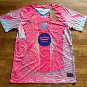 Barcelona Fotbollströja - Säljer en fet FC Barcelona x Nike fotbollströja i rosa och ljusblått med coola grafiska linjer och Spotify-logga på bröstet. Tröjan har korta ärmar, V-ringning och är gjord i lätt, ventilerande material. Perfekt för dig som vill sticka ut på planen eller läktaren.