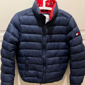 Mörkblå dunjacka Tommy Hilfiger - Snygg mörkblå dunjacka från Tommy Hilfiger med röd insida och klassisk logga på ärmen. Jackan är i dun med dragkedja framtill och två fickor med dragkedja. 