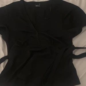Svart omlottopp från Gina Tricot XS - Säljer en svart omlottopp från Gina Tricot i storlek XS. Toppen har korta ärmar, v-ringning och knytband i midjan för en snygg passform. Aldrig använd perfekt för middag med familj ut osv.