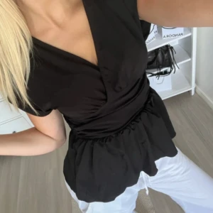 Svart omlottopp med volang - Säljer en svart omlottopp med v-ringning och korta ärmar. Toppen har markerad midja och volang nedtill som ger en snygg siluett. Perfekt att styla med både jeans och kjol. Size S🖤