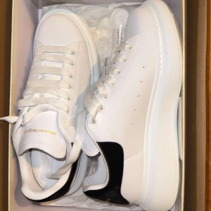Alexander McQueen vita sneakers - Säljer ett par klassiska vita Alexander McQueen sneakers med svart detalj på hälen och tjock, platt sula. Skorna har snörning och är tillverkade i skinn med diskret guldtext på plösen. Perfekta för dig som gillar stilrena och trendiga sneakers.