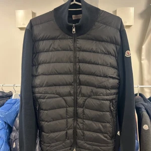Svart cardigan jacka från Moncler - Svart cardigan jacka från Moncler med quiltad framsida i dun och stickade ärmar och rygg. Jackan har dragkedja framtill, ribbad krage och Moncler-logga på ärmen. Perfekt för dig som vill ha en stilren och sportig look.