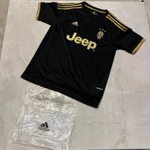 Juventus svart guld fotbollströja Adidas - Juventus matchtröja från Adidas i svart med guldiga detaljer och tryck. Klassisk rund hals, korta ärmar och tre guldiga ränder på axlarna. Jeep-logga på bröstet och Juventus klubbmärke. Tillverkad i lätt och ventilerande funktionsmaterial.