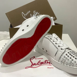 Christian Louboutin vita sneakers med nitar - Snygga vita sneakers från Christian Louboutin med ikonisk röd sula. Skorna är i skinn och har silverfärgade nitar på tå och häl för en edgy look. Klassisk snörning och rund tå. Perfekta för dig som vill sticka ut med exklusiv stil.