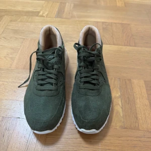 Nike Air Max 90 i mörkgrön mocka - Snygga Nike Air Max sneakers i mörkgrön mocka med vita sulor och beige detaljer vid hälen och insidan. Klassisk siluett med snörning och rund tå, perfekt för dig som gillar streetstyle och vill sticka ut med en unik färg. Skorna har en platt sula och ikonisk Air Max-dämpning.