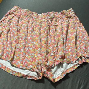 Blommiga shorts i bomull - Säljer ett par superbekväma shorts med blommigt mönster i rosa, blått och gult på en orange bas. De har elastisk midja och är perfekta för varma dagar. Lätta och luftiga i mjuk bomull, passar perfekt till sommaren.