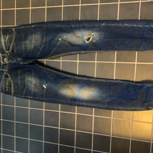 Blå jeans från Dondup med slitningar - Säljer ett par blå jeans från Dondup med coola slitningar och hål på låret. Jeansen har klassisk femficksdesign, kontrastsömmar och en snygg tvättad look. Tillverkade i jeansmaterial och har en rak passform som sitter snyggt.