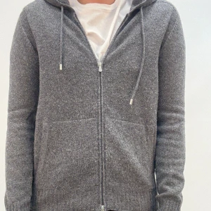 Grå stickad hoodie med dragkedja - Mysig grå stickad hoodie med dragkedja och huva. Tröjan har långa ärmar, ribbade muddar och två fickor framtill. Perfekt för kyliga dagar och enkel att matcha med det mesta. Snygg och stilren design med snörning i huvan.