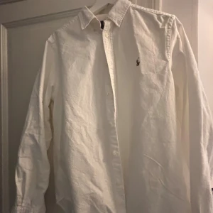 Vit skjorta från Ralph Lauren - Klassisk vit skjorta från Ralph Lauren med broderad logga på bröstet. Skjortan har lång ärm, knappar framtill och en snygg krage. Tillverkad i mjuk bomull som känns skön mot huden. Perfekt för en clean och stilren look. 1600kr