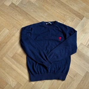Mörkblå tröja från Ami paris  - Mörkblå långärmad tröja från Ami Alexandre Mattiussi med rund hals och röd broderad hjärtlogga på bröstet. Tröjan är ribbad vid ärmslut och nederkant och har en clean, klassisk look som funkar till allt.