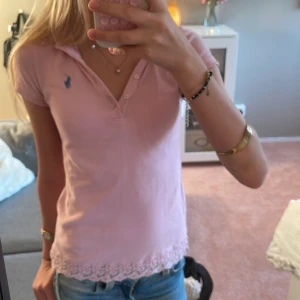 Rosa pikétopp från Ralph Lauren - Söt ljusrosa pikétopp från Ralph Lauren med klassisk krage, korta ärmar och knappar framtill. Liten blå logga på bröstet och spetsdetalj längs nederkanten som ger en extra feminin touch. Perfekt till jeans eller kjol.