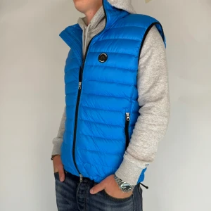 J.Lindeberg Dunväst - Nu har vi nöjet att erbjuda en exklusiv Dunväst från det välrenommerade märket J.Lindeberg. Dunvästen är i storlek Large och är i mycket gott skick utan några synliga defekter. Modellen på bilden är 180 cm lång och väger 75 kg, vilket kan ge en uppfattning om passformen