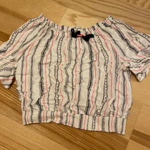 Randig croppad topp från H&M - Säljer en croppad topp från H&M med elastisk halsringning och nederkant. Toppen är vit med svarta och rosa ränder i olika mönster och har en söt knytning med tofsar framtill. 