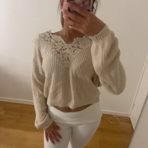 Beige stickad tröja med spetsdetalj - Superfin beige stickad tröja med långärmade ärmar och croppad passform. Tröjan har en snygg spetsdetalj i vitt vid halsen som ger en extra touch. Perfekt för dig som gillar mjuka material och vill ha något som sticker ut lite.