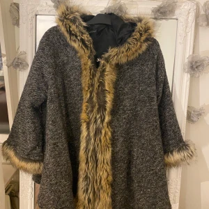 Grå kappa med fuskpälsdetaljer - Säljer en snygg grå kappa med huva och breda ärmar, dekorerad med mjuk fuskpäls i bruna toner längs huva, ärmslut och framkant. Kappan har ett grovt vävt tyg och en loose passform, perfekt för kalla dagar när du vill vara både varm och stilren.