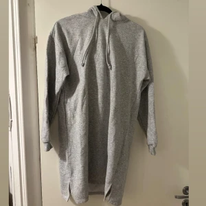 Grå hoodieklänning med huva - Mysig grå hoodieklänning med långa ärmar, huva och snörning framtill. Klänningen har en avslappnad passform och är tillverkad i mjuk bomullsmix. Perfekt för chill dagar och enkel att styla med sneakers eller boots.