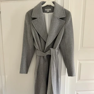 Grå kappa med bälte från H&M - Stilren oanvänd grå kappa från H&M med bred krage och knytskärp i midjan. Lång modell med klassisk passform och långa ärmar. Går ned över knäna. Perfekt för lager-på-lager och snygg till både jeans och boots. Gjord i mjukt ullblandat material för en lyxig känsla. 