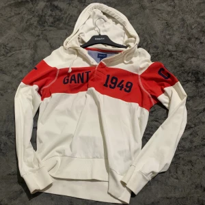 Vit och röd hoodie från Gant - Snygg hoodie från Gant i vitt med breda röda partier och blå textdetaljer. Tröjan har huva med snörning, långa ärmar och ribbade muddar. Perfekt för dig som gillar sportig och avslappnad stil.