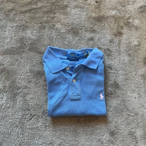 Blå Polo Ralph Lauren pike  - Snygg blå piké  från Polo Ralph Lauren med klassisk pikékrage och två knappar framtill. Den har korta ärmar och en broderad rosa logga på bröstet. Tillverkad i mjuk bomull som är skön mot huden. Perfekt för en chill och stilren look.