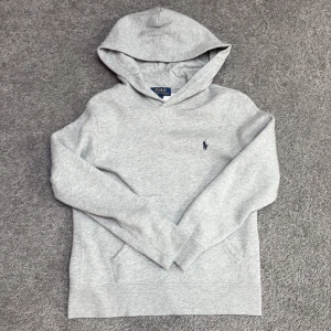 Grå hoodie från Polo Ralph Lauren - Snygg ljusgrå hoodie från Polo Ralph Lauren med klassisk huva och broderad marinblå logga på bröstet. Tröjan har känguruficka framtill och är tillverkad i mjuk bomull. Perfekt för chill dagar och enkel att matcha med jeans eller joggers.